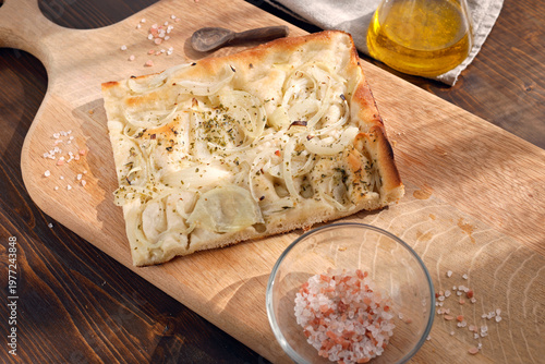 Traditional Italian onion focaccia on wooden cutting board with pink salt. High-angle shot features aromatic herbs olive oil and rustic kitchen background under warm natural sunlight. Professional