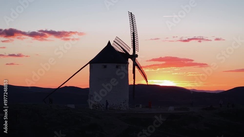 Traditional windmill from Consuegra, Spain, turning its blades during a beautiful orange sunset with mountains in the background and a plane flying in the sky