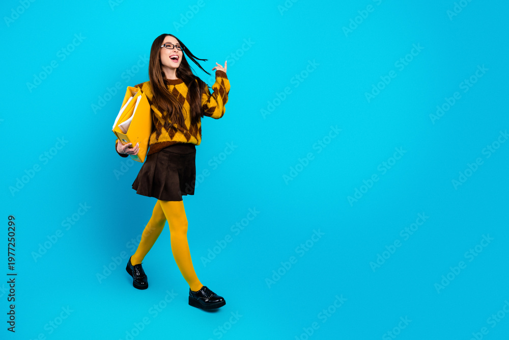 Fototapeta premium Happy young student walking with yellow folder and tights on blue background wearing argyle sweater and glasses, stylish schoolgirl portrait full length with confident joyful expression