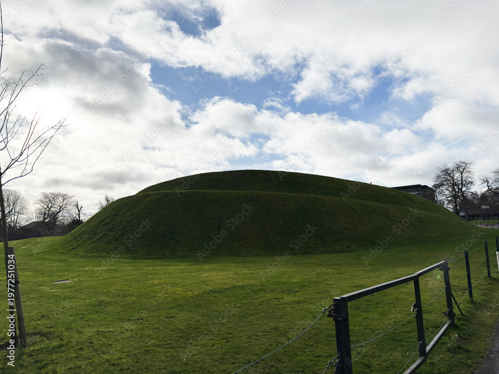 Fototapeta premium Grassy earthwork mound and green hill in a park with dramatic cloudy sky in Edinburgh, Scotland, UK historic landscape scene
