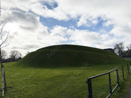 Grassy earthwork mound and green hill in a park with dramatic cloudy sky in Edinburgh, Scotland, UK historic landscape scene