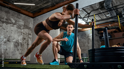 Woman pushing weighted sled while man supervising training session in gym. Concept of strength training promo, fitness ads campaign, athletic branding and performance coaching service marketing
