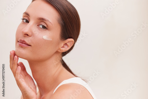 Canvas Print Anti-aging skincare woman actively blending face cream on her cheekbone with fingertips while looking directly at the camera, with copy space on a soft neutral background