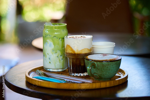 Tray with clay mug of cappuccino, glass latte, and matcha latte on small wooden table in Vietnamese coffee shop, beautiful bokeh background.