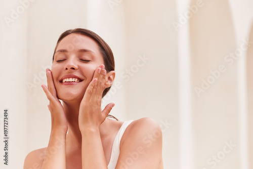 Canvas Print Facial care woman smiling joyfully with both hands cupping her cheeks and eyes closed, glowing bare skin highlighted against a soft Mediterranean-style column background with copy space
