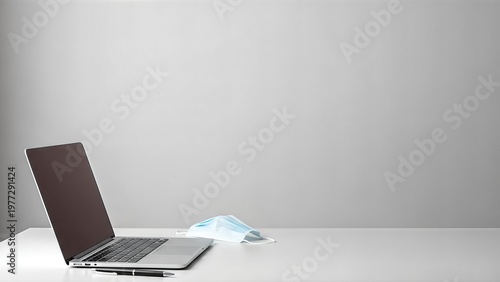 A laptop computer and a face mask are placed on a white desk, symbolizing remote work and health precautions.