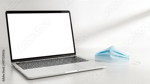 A modern laptop with a blank screen sits next to a blue surgical face mask on a white desk, symbolizing remote work and health precautions.