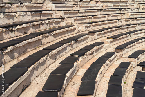 Stone amphitheater seating with rows of steps and black cushions arranged in curved tiers, outdoor venue for audience and events. Amphitheater seating usage for event venue, performance space