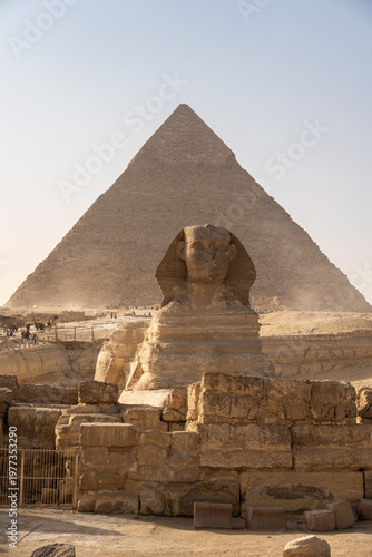 A majestic front-facing view of the Great Sphinx of Giza rising from the desert plateau, with the towering Pyramid of Khafre dominating the background beneath a hazy golden sky.