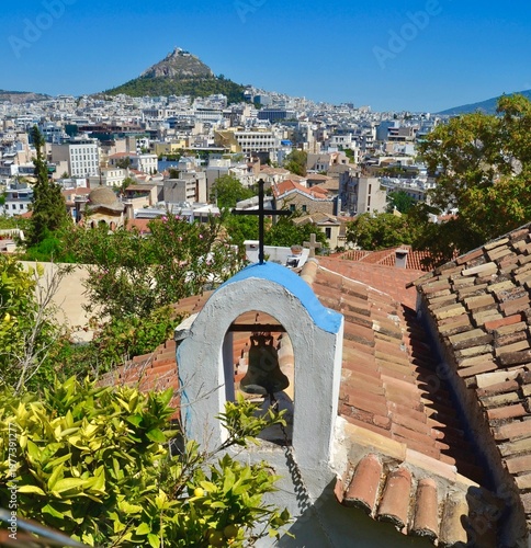 Cityscape of Athens, Greece, with a focus on a small church bell structure featuring a cross. . In the background, the densely built urban landscape of Athens  and landmark  Lycabettus  hill