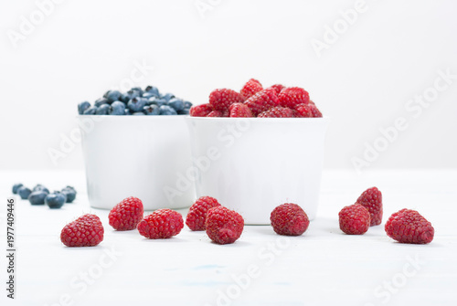 bilberry and raspberry fruits at white ceramic cup, on bright wood table