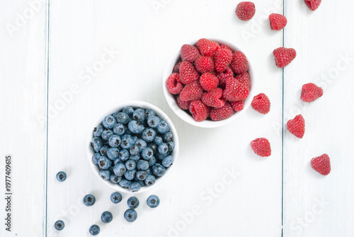 bilberry and raspberry fruits at white ceramic cup, on bright wood table