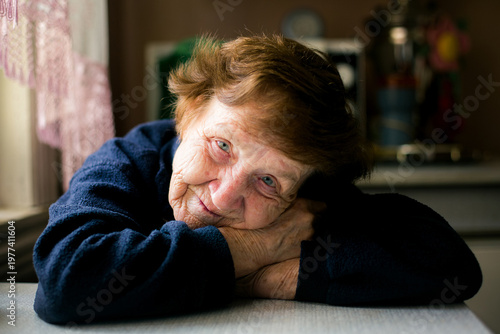 Portrait of elderly woman resting her head on hands by a table near window with soft natural light and warm home interior.