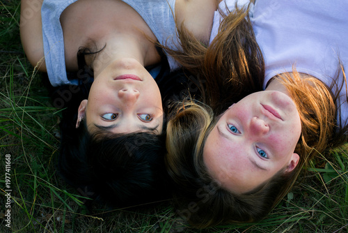 Two young girls lying on grass outdoors, looking at each other in natural light, top view lifestyle moment of friendship and connection.