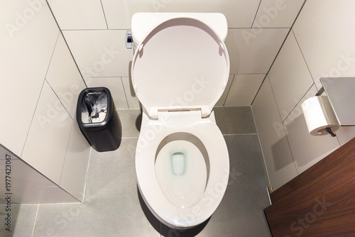 White ceramic toilet with open lid stands on gray tiled floor. Black trash bin with plastic liner placed beside it. Toilet paper holder mounted on wall in modern bathroom.