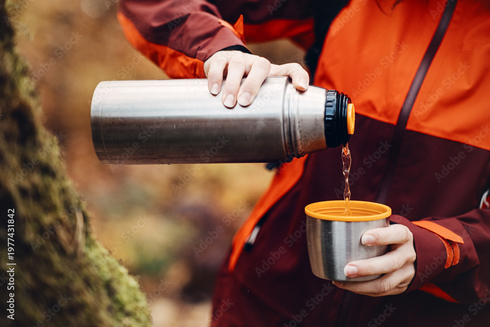 Naklejka premium Hands pour hot liquid from thermos into cup with smiling woman in background. tea break outdoors, coffee on hike, hot drink in forest, warming up outdoors.