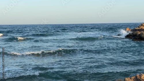Ocean waves crashing on rocky shore under clear sky, ideal for coastal background images