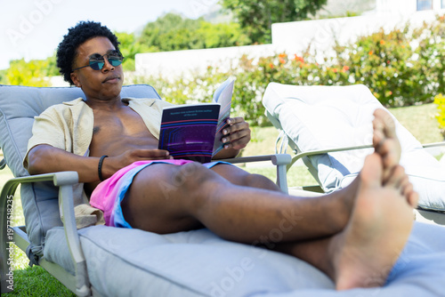 African American man reclining on lounge chair in backyard, in pink trunks reading purple book