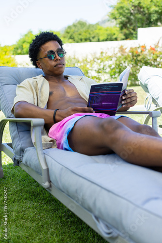 Adult African American man reclining on lounger in backyard wearing swim shorts reading paperback