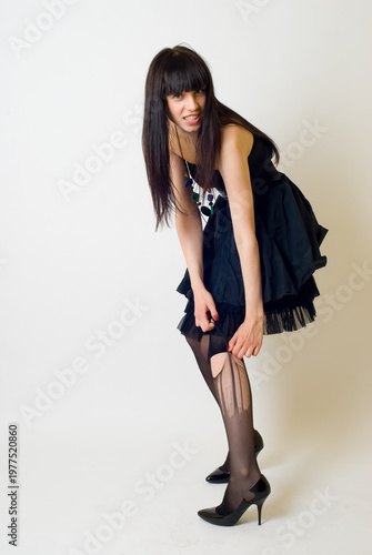 Studio portrait of a young woman wearing an elegant black dress, posing against a clean white background. The model is captured in a full-length or three-quarter pose, exuding confidence