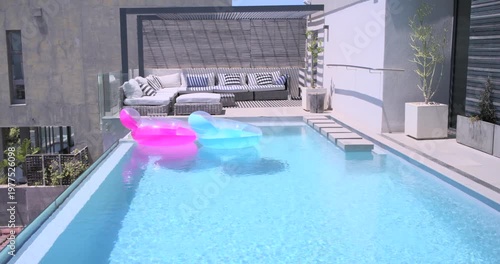 Two inflatable floats drifting across rooftop pool under gentle breeze, showing pergola and sofa