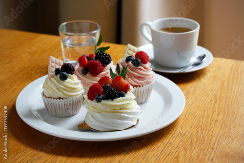 Assorted cupcakes with fresh berries and cream on white plate with coffee and water on wooden table, cozy cafe dessert scene, sweet pastry selection for brunch or afternoon treat