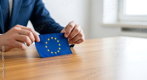 Businessman in a professional navy blue suit holding a small European Union flag on a wooden office desk, representing political agreements and diplomacy. Realistic photography with selective focus.