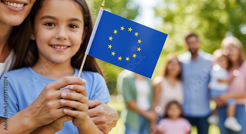 Smiling young girl holding a small European Union flag during a sunny family picnic in a park with blurred background people. Lifestyle portrait photography with natural sunlight and bokeh.