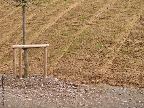 Medium shot of a sloped embankment covered with brown coir netting for
erosion control. The biodegradable geotextile mesh stabilizes the hillside while
young plants grow through the grid. 