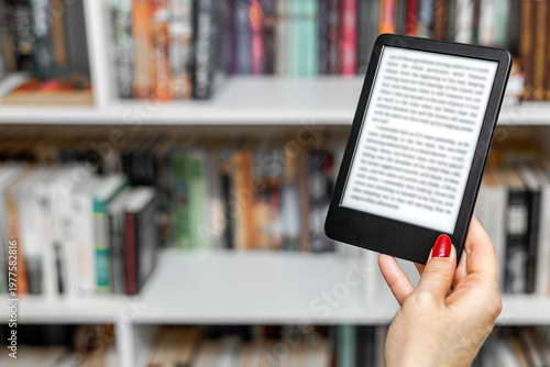 a woman's hand holding an e-book. A woman reads an e-reader near a bookcase. an e-book against the background of paper books. A woman reading an e-book at home. isolate of a hand holding an e-book