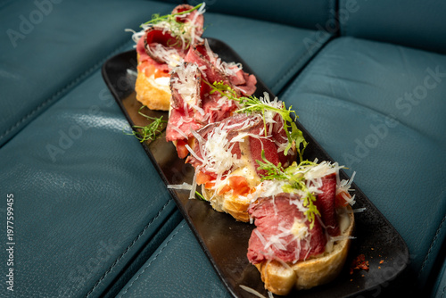 Four bruschettas with roast beef, parmesan, and microgreens on a black plate against a dark teal leather background.