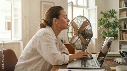 Professional woman suffering from intense summer heat, finding refreshing comfort, enjoying the cooling breeze of an electric fan, working at her home office during a sweltering day