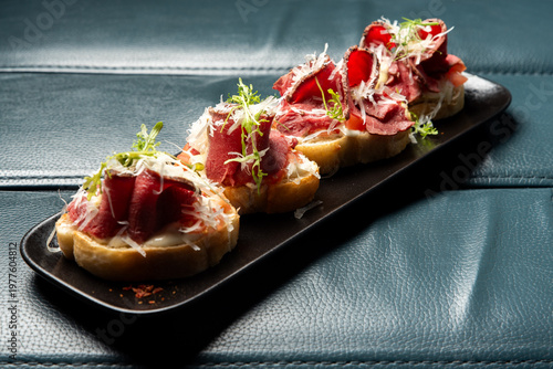 Beef carpaccio bruschettas with parmesan and greens on a black platter, set on a blue leather surface.