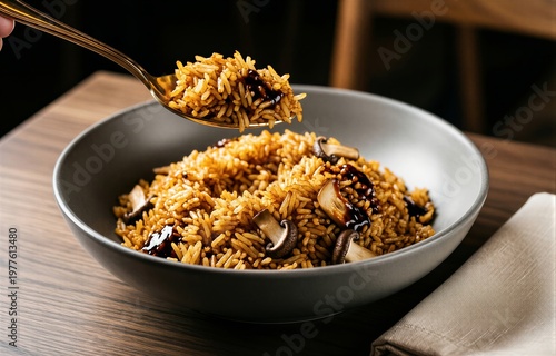 Bowl of Nasi Goreng with sliced mushrooms and dark sauce drizzled over it. Concept of fried rice with sweet soy served indoors, warm lighting enhances cozy atmosphere.