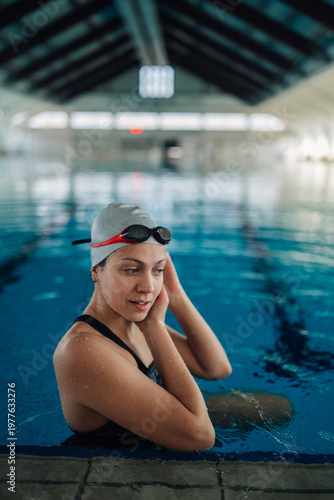 Obraz na plátne Young woman swimmer with goggles and swim cap by the pool edge in an indoor stad