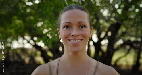 Portrait of pretty woman in sports bra smiles as stands against green tree closeup. Cheerful lady with daily makeup expresses positive walking in summer park