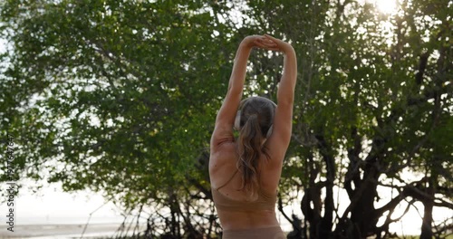Young woman with headphones warms up body before yoga training in park rear view. Flexible sportswoman stretches back and arms standing among summer nature