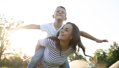 Smiling mother giving her son a piggyback ride outdoors, playful family moment, child pretending to fly, summer fun and Mother’s Day concept.
