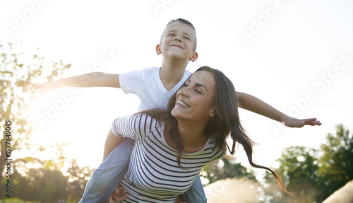 Smiling mother giving her son a piggyback ride outdoors, playful family moment, child pretending to fly, summer fun and Mother’s Day concept.
