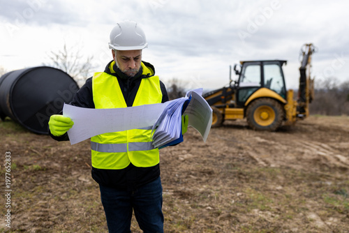 Vászonkép Engineer reading construction documents with backhoe on site