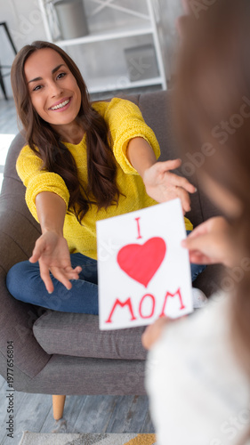 Happy mother reaching out to receive a handmade Mother’s Day card from her daughter at home. Family love, bonding, care, appreciation and holiday lifestyle concept.