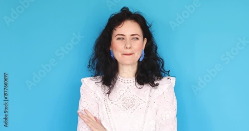 Video of happy coy young woman in white lace blouse hugging herself and then playfully shrugging with smile isolated on blue background, expressive female portrait of joy and flirtation