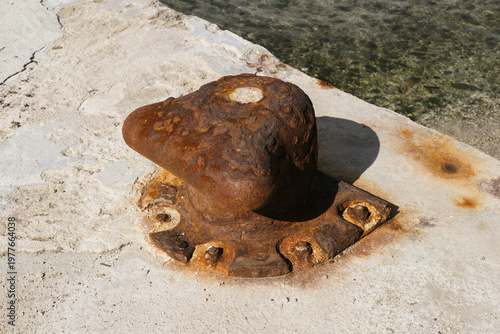 Rusty mooring bollard on concrete pier by the water, weathered metal, marine environment, nautical element, industrial design, coastal scenery.
