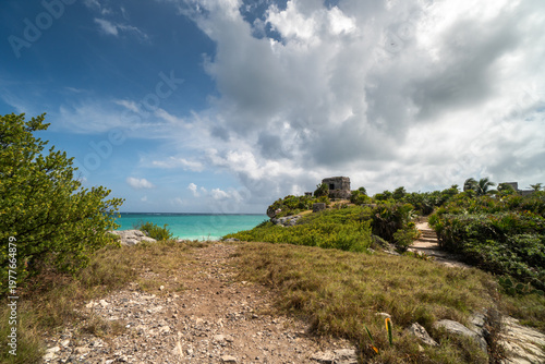 Scenic view of ancient ruins overlooking the turquoise sea in Tulum Mexico. Lush greenery and dramatic clouds enhance the landscape.