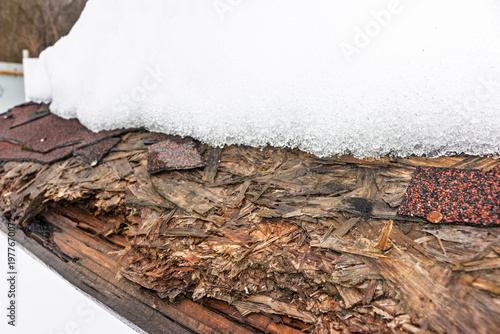 Fotografie Melting snow on a damaged asphalt shingle roof edge, revealing rotten plywood and weathered wood structure