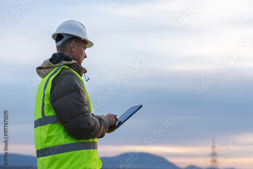 A young male electrical engineer works outdoors, inspecting high-voltage power lines with a tablet in a utility area at dusk.
