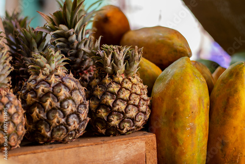 A close-up of exotic fruits—pineapples and papayas—lies on a wooden surface. Several ripe pineapples are in the foreground. The background is blurred, with selective focus. Fruit sales at a market.