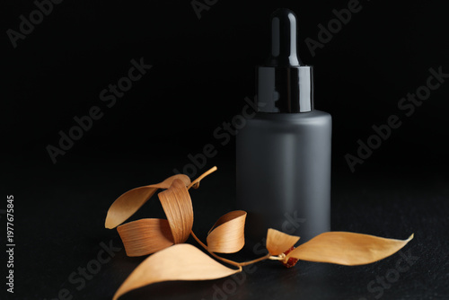 Bottle of cosmetic product and dry leaves on dark textured table against black background