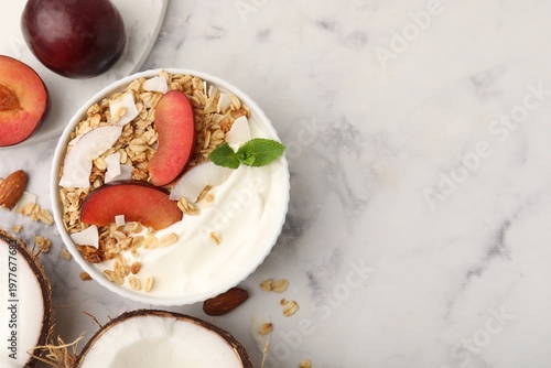 Delicious coconut yogurt with granola and plum in bowl on light marble table, flat lay. Space for text