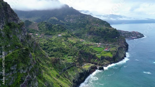Aerial view of Madeira's dramatic coastline, featuring lush terraced hillsides, Boaventura villages, and the vibrant Atlantic Ocean under a cloudy sky.